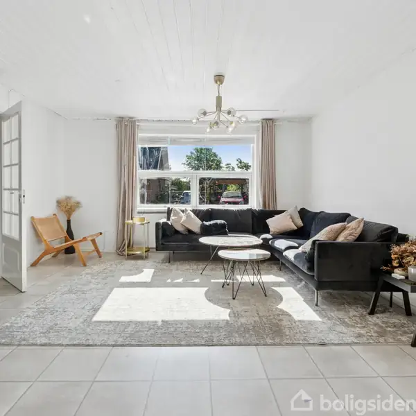 Stue med sort sofa arrangeret i en L-form og sofabord foran et stort vindue. Lyse vægge og gulv, en stol til venstre, plante på sidebord, moderne loftslampe.