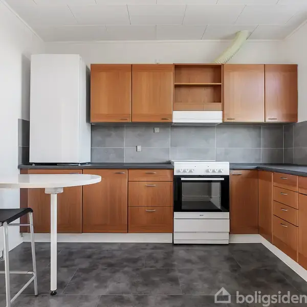 Et moderne køkken med træskabe, grå fliser og en hvid ovn. Et sort og hvidt barstol ved et rundt bord. Lys strømmer ind fra et vindue til højre.