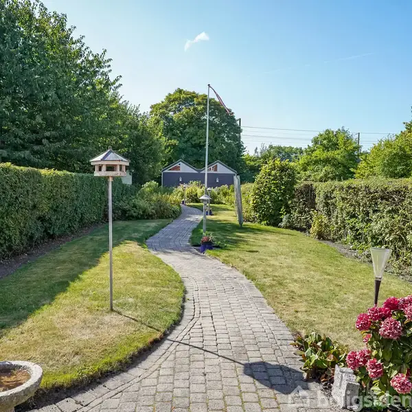 En snoet brostenssti løber gennem en velplejet have med buske og blomster langs kanten. Fuglehuse står på pæle, og en flagstang står i baggrunden foran en hæk.