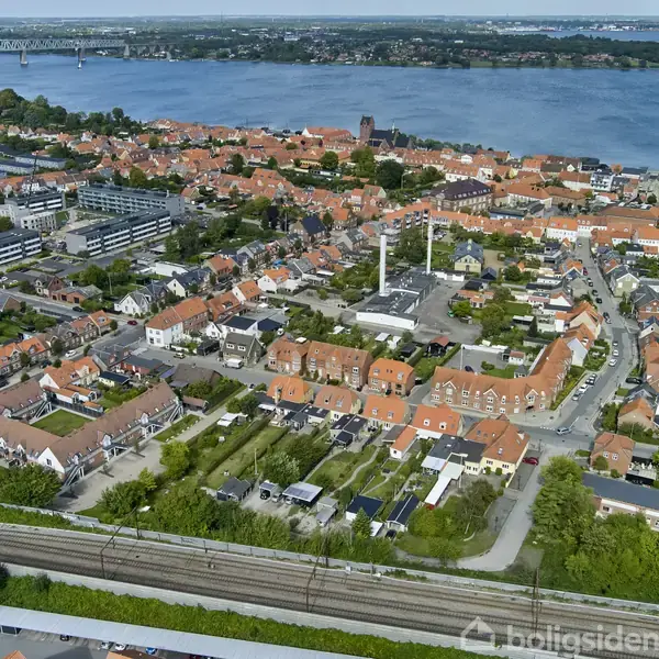 By i fugleperspektiv med røde tage og grønne områder, jernbanespor i forgrunden, flere industriskorstene, en flod i baggrunden med en bro, omgivet af spredte bygninger og beboelseshuse.