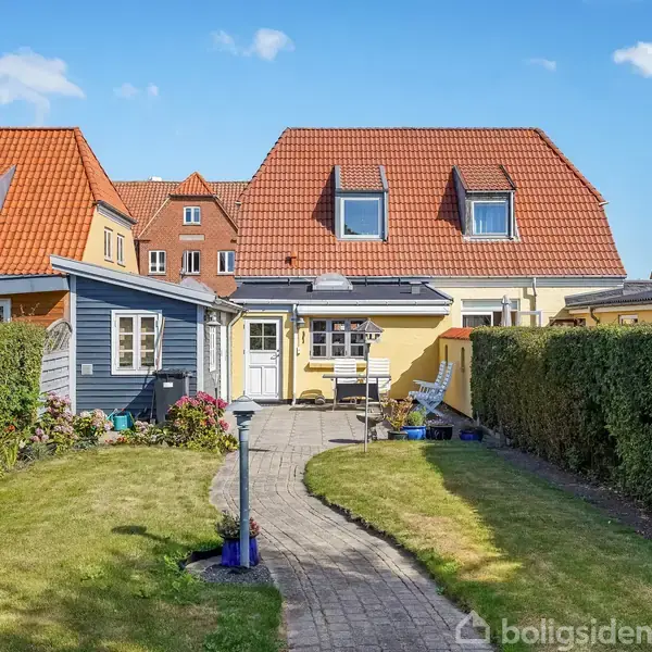 Et lille hus med rødt tag ses i baggrunden af en velplejet have. En stensti fører gennem haven, flankeret af grønne hække og græsplæner.