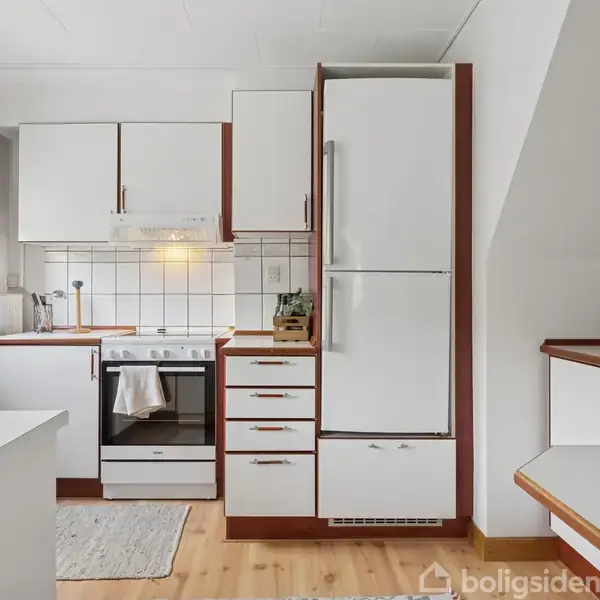 Et funktionelt køkken med hvide skabe og en ovn i midten. Et køleskab står til højre. Tæpper ligger på det lyse trægulv. Hylder med skåle er til højre.