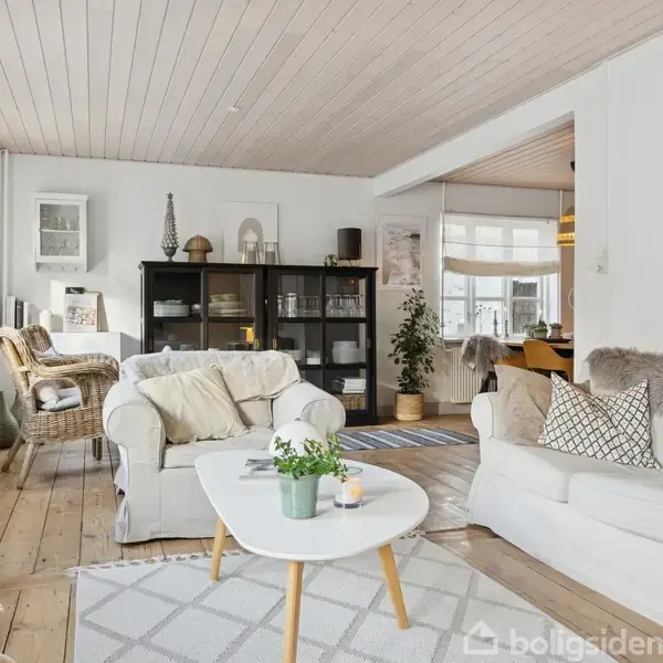 En lys stue med hvide møbler og træmøbler. Et sofabord har planter på sig. Rummet har lyse trægulve og store vinduer med persienner, hvilket skaber en hyggelig atmosfære.