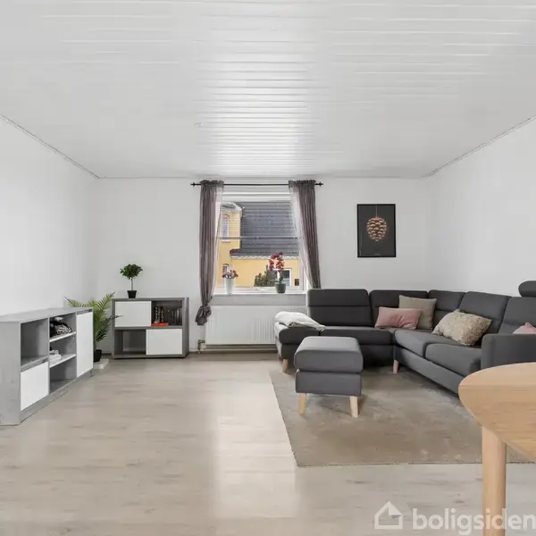 Stue med en stor grå sofa og skammel på et tæppe, ved siden af et rundt træbord. Et vindue med gardiner slipper lys ind, og en vægplakat hænger ved sofaen.