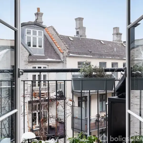 Åben altandør med udsigt til grå boligbygninger. En blomsterkasse med planter hænger på gelænderet. Kviste og skorstene ses på tagene i en tæt bymæssig bebyggelse.