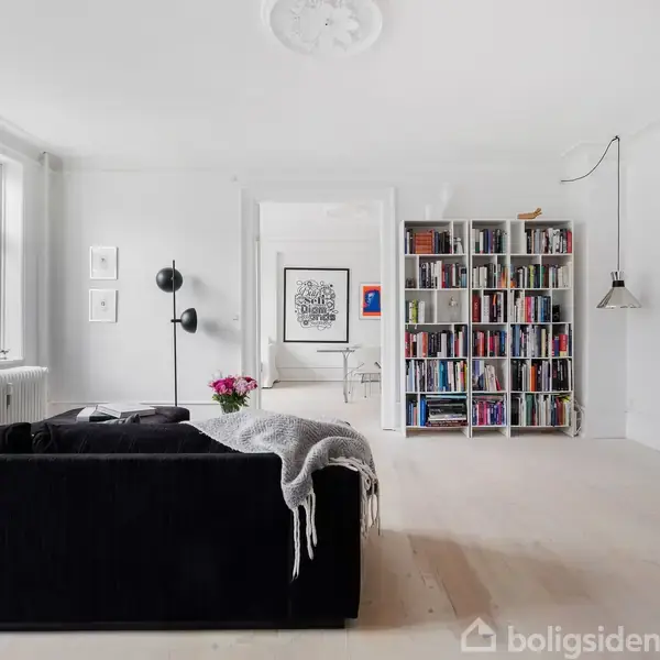 Sort sofa står i en lys stue, med en reol fyldt med bøger og moderne kunst på væggene. Store vinduer lyser rummet op. Pejsen er omringet af stole.