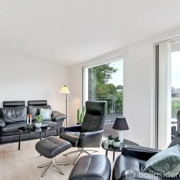 Sort læderlænestol og sofa står i en stue med hvidvægge. Stuen har store vinduer med udsigt til have og huse. Lampe og planter tilføjer hygge.