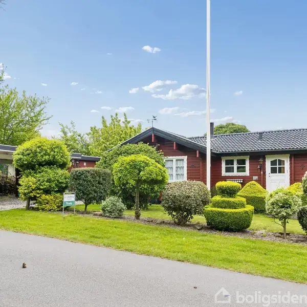 Et rødt hus står stille omgivet af en velplejet have med trimmede buske, ved siden af en carport. Det er dag, og himlen er blå med spredte skyer.