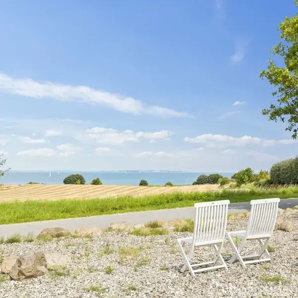 To hvide stole står på en grusbelagt terrasse, hvor de vender ud mod en kornmark og kyststrækning med havudsigt under en blå himmel med spredte skyer. Træer rammer scenen.