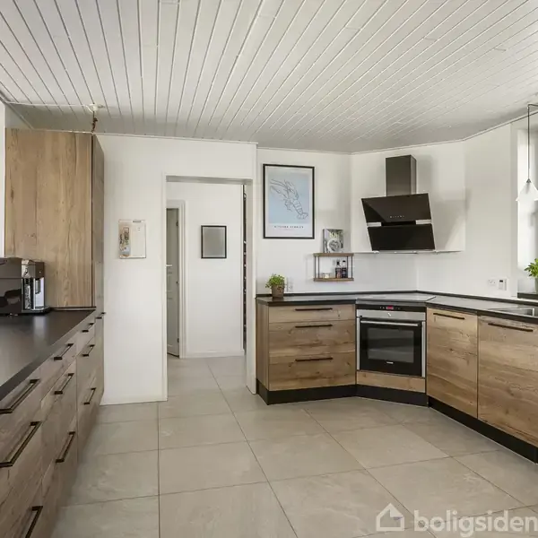 Køkken med træskabe og sorte bordplader. Kaffemaskine på venstre bord, ovn og kogeplade centreret. Lysindfald fra stort vindue til højre, loft med paneler. Ingen tekst til stede.