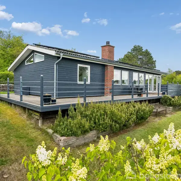 Et moderne, blåmalet hus med store vinduespartier står på en hævet terrasse omgivet af en grøn have med buske og træer under en skyfri himmel.