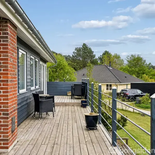 Veranda med trædækning set fra hus med murstens- og træbeklædning. To stole og et bord står mod en balustrade. Omgivende huse og træer i grønne omgivelser.