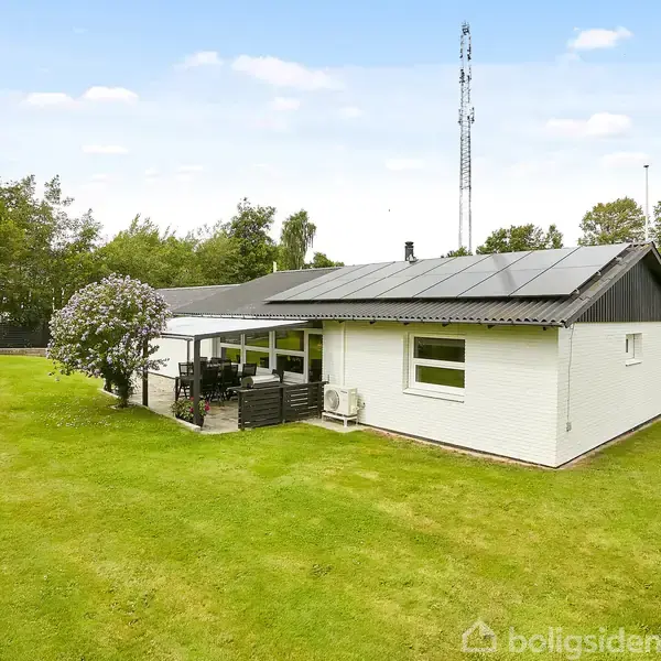 Et hvidt hus med solpaneler på taget står stille i en grøn have. Der er et overdækket terrasseområde med havemøbler. Omkring huset er der græsplæne og træer.