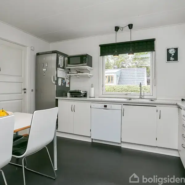 Et lyst køkken indeholder et spisebord med frugtskål og fire stole. Et vindue med udsigt til have ses over en vask. Køleskabet står ved siden af en mikrobølgeovn.