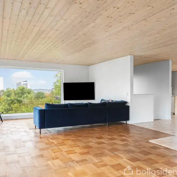 Brændeovn opvarmer stue, hvor en blå sofa og fladskærms-TV vender mod panoramaudsigt af grønne træer og en by i det fjerne. Lys træloft og parketgulv skaber en hyggelig atmosfære.