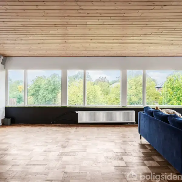 Stue med blå sofa der står mod en væg af store vinduer. Udsigt til grønne træer udenfor. Trægulv og træloft skaber en lys og åben atmosfære.