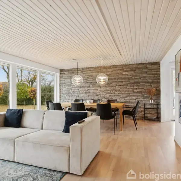 En beige sofa står foran store vinduer, der viser en have udenfor. Spisebord og stole er opstillet ved en murstensvæg under loftslamper. Væggene har kunstplakater.