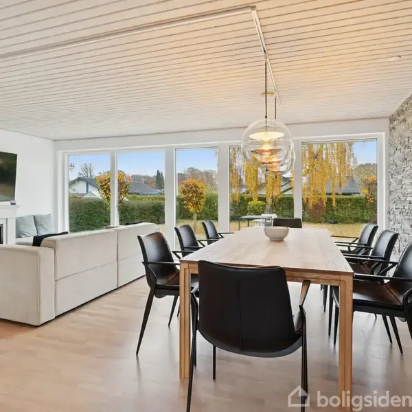 Stue med hvid sofa og væghængt TV over pejs, ved siden af spisebord med sorte stole. Store vinduer viser have udenfor. Væg dekoreret med rå mursten.