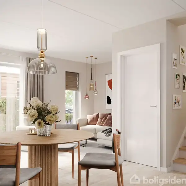 Rundt spisebord med blomsterbuketter står i lys stue. Moderne møblering med sofa, stor vinduesfacade, lyse vægge og trapper til venstre. Flere billeder hænger langs trappen.