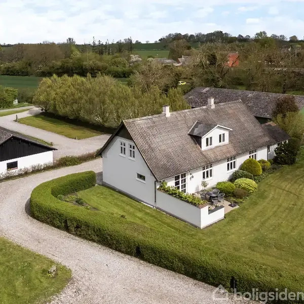 Luftfoto af en gård med hvidmalede bygninger og stråtækte tage, omkranset af grønne marker. En indkørsel fører op til gården, omgivet af velplejet have og enkeltstående træer.