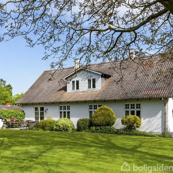 Et hvidt hus med rødt tag står stille i en frodig have. Buske omkranser huset, og en terrasse med havemøbler ligger til venstre. Sollys oplyser scenen.