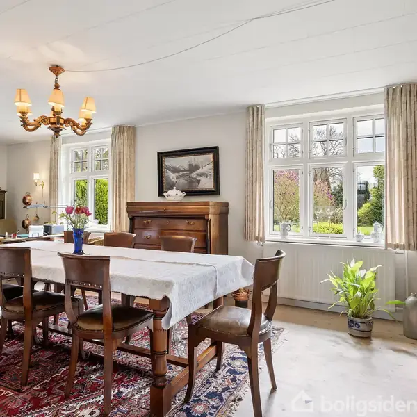 Et hvidt hus med gråt tag ligger omgivet af en velplejet have og indkørsel. Huset har en fliseterrasse, og rundt om ses marker og træer under en blå himmel.