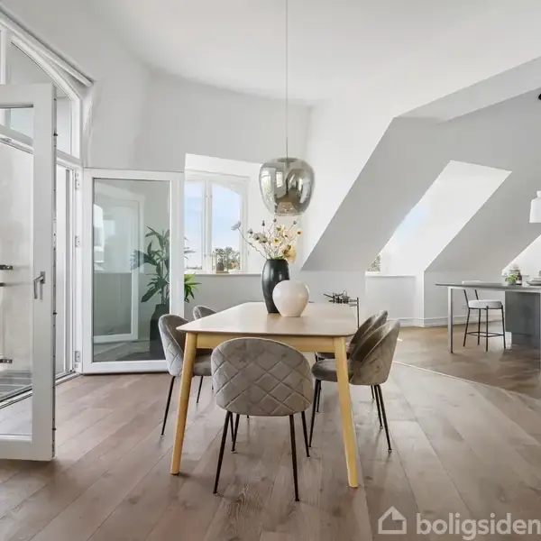 Spisebord med vase og stole står i en lys stue med åbne franske døre. Bagved ses et moderne køkken med barstole og hængelamper, alt omgivet af store skråvinduer.
