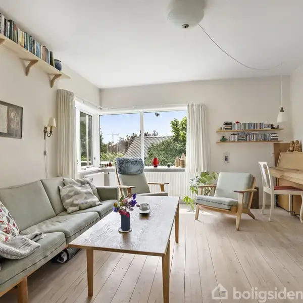 Stue med lysegrå sofa, sofabord med vase, og en lænestol. Et klaver står ved væggen, og hylder med bøger er placeret over sofaen. Store vinduer giver udsigt til grønne træer.