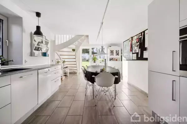 Et moderne køkken-alrum med hvidt interiør, sorte stole omkring et bord. Trappen fører til en anden etage. Store vinduer giver udsigt til have. Fliser på gulvet.