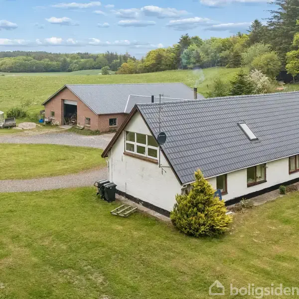 Hvidt hus med sort tag står på en grøn græsplæne. I baggrunden ses en rød bygning og skov, under en blå himmel med spredte skyer.