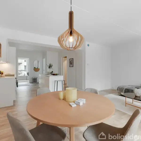 Rundt spisebord med stole, omgivet af minimalistisk indrettet stue med sofa og sofabord. En moderne lampe hænger over bordet. Åbent køkken ses i baggrunden. Billeder på væggen.