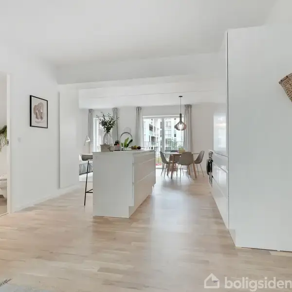 Køkkenø i et lyst rum med trægulv, omgivet af spisebord og stole ved store vinduer. En dør til badeværelse til venstre, knagerække med kurv og jakke på væggen.