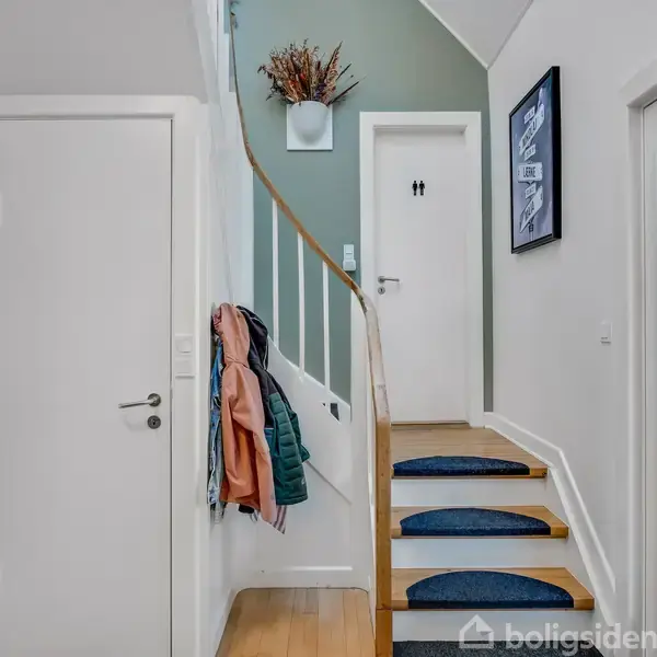 Trappeopgang fører opad med tæppebelagte trin. Jakker hænger på knager ved siden af hvide døre. På væggen hænger en plakat og en vase med grene, der skaber en hjemlig atmosfære.