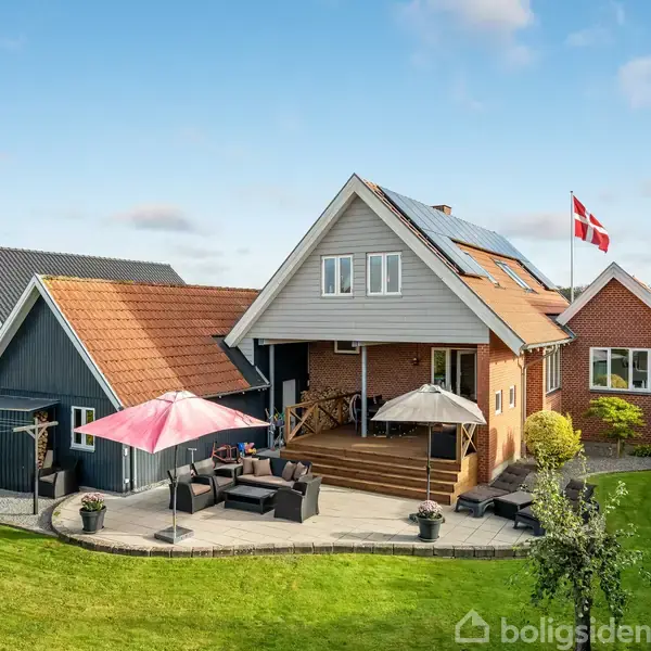 Et rødt murstenshus med flagstang står i en velplejet have. På terrassen er der loungemøbler og paraplyer. Huset har solpaneler på taget og naboen er tæt på.