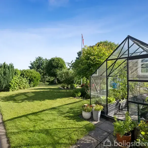 Et drivhus står i en frodig have, omgivet af forskellige planter og en græsplæne. Huset ses på venstre side med en flisebelagt terrasse foran. Himlen er klar og blå.