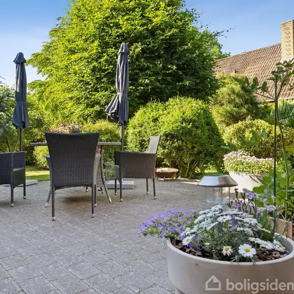 Møbleret terrasse med borde og stole er omkranset af frodig have med buske og træer. Blomster i krukker dekorerer området. Solsikkeparasoller står midt i det grønne miljø.