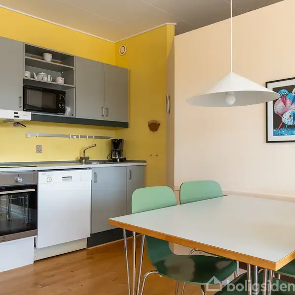 Køkken med grå skabe og en ovn ved siden af en opvaskemaskine. Et spisebord med grønne stole står i forgrunden. Gule vægge og en lampe hænger ned fra loftet.