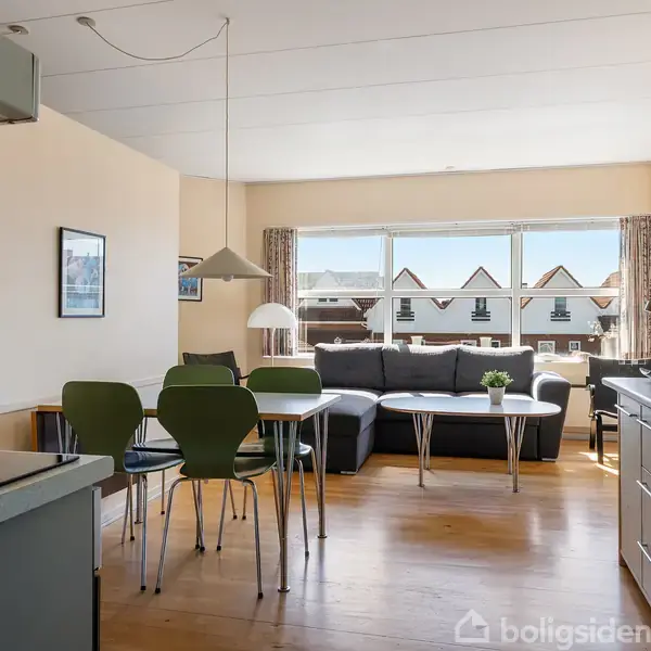 Stue med træbjælker har en grå sofa ved store vinduer. Spisebord med grønne stole står centralt. Køkkenet ses til venstre. Billedramme hænger på væggen.