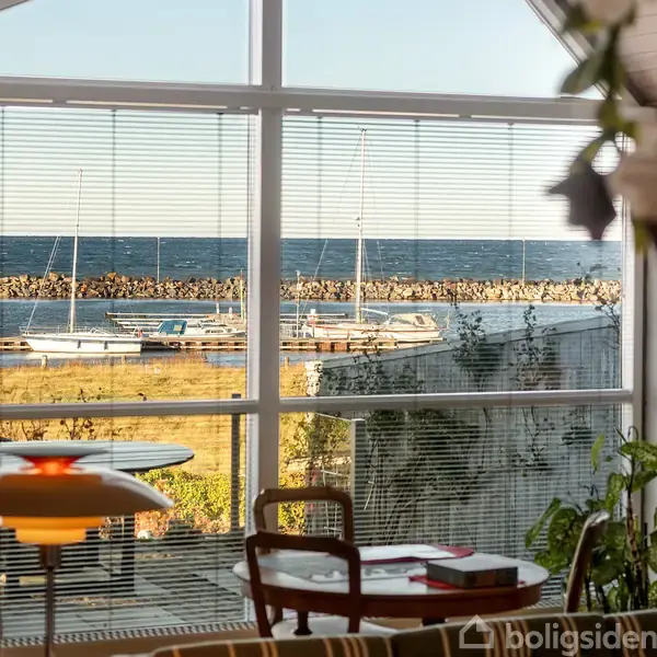 Indendørs med udsigt over havet, ses sejl både i marinaen gennem store vinduer med persienner, omgivet af moderne møbler, grønne planter og belysning.