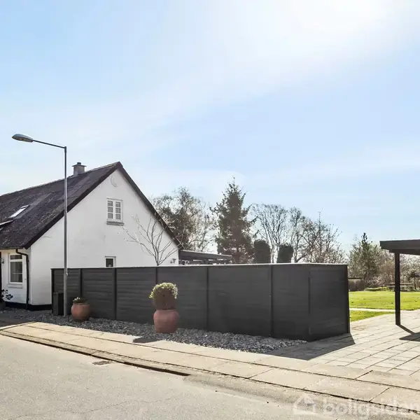 Hvidt hus med sort tag står ved siden af en sort hegn. I forgrunden er der en asfalteret vej og en garage. Træer i baggrunden skaber et landligt miljø.