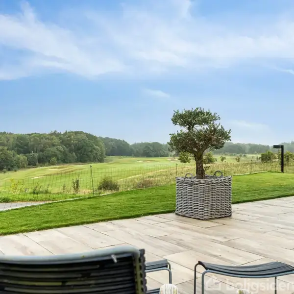 Stole står på en terrasse, der vender ud mod en grøn plæne og en golfbane. En lille træ i potte står ved siden af stolene. Himlen er delvist skyet.