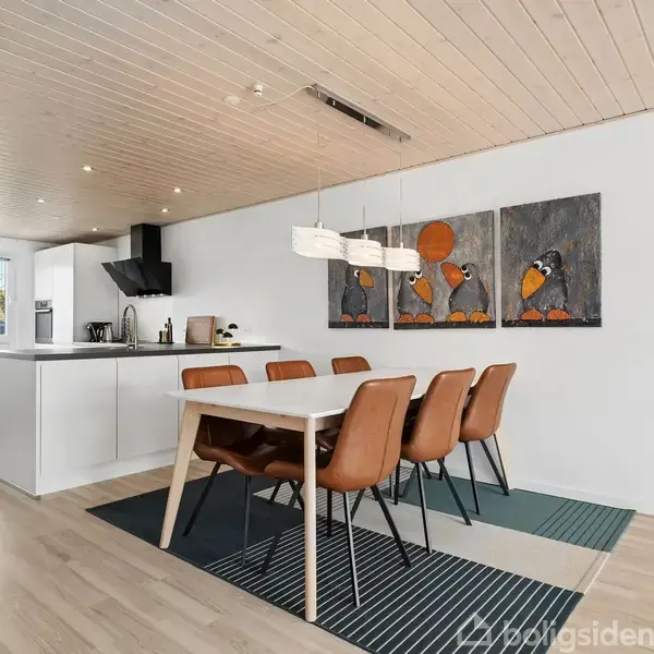 Spisebord med seks brune stole står på tæpper. Billedkunst pryder væggen. Til venstre er et moderne køkken med hvide skabe, under et lyst træloft.