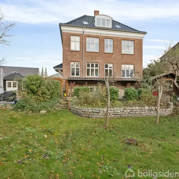 En stor, treetagers rødstensbygning står stille omgivet af en frodig have med græs og buske. En stensat terrasse ses foran huset. Himlen er blå med lette skyer.