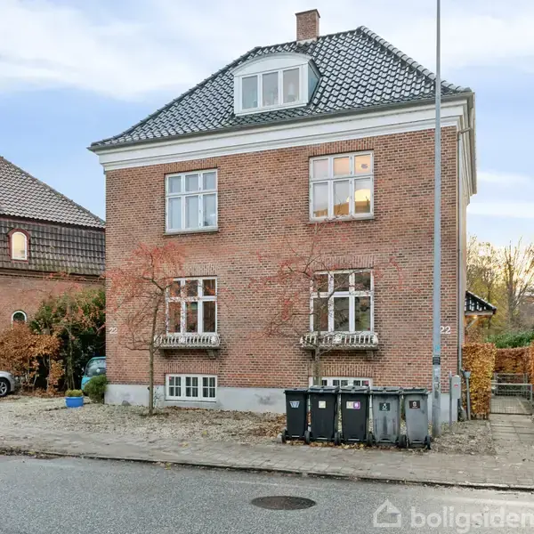 Murstenshus med tre etager og sort tegltag, står på gadehjørne ved siden af parkerede biler og skraldespande. Huset har hvidrammede vinduer og er omgivet af efterårsløv.