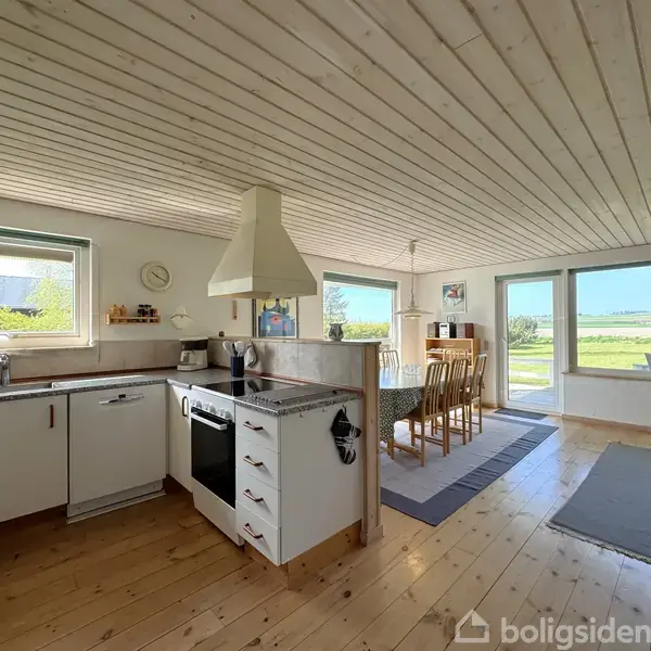 Et lyst køkken-alrum med hvide skabe og en ovn er åbent mod en spiseplads med seks stole. Store vinduer og terrassedøre giver udsigt til en grøn have.