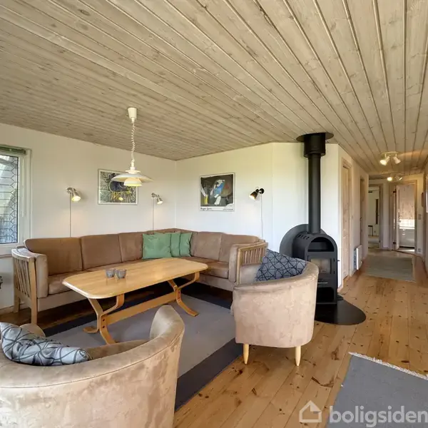 Stue med en brun hjørnesofa og to beige lænestole omkring et træsofabord, placeret ved siden af en sort brændeovn. Lyse trævægge og loft, to billeder hænger på væggen.