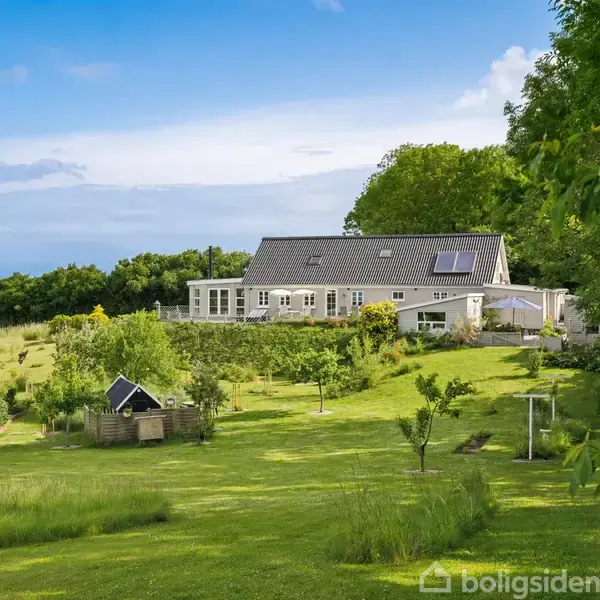 Hus med solpaneler står stille i en frodig have med træer og græs, omgivet af skov og blå himmel i baggrunden.