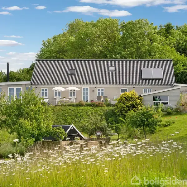 Hus på landet med mørkt tag og flere vinduer, omgivet af grønne træer og en blomstereng i forgrunden. Solpaneler er synlige på taget.