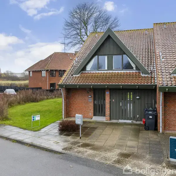 En halvanden plans hus med rødt tegltag står i et villakvarter. En græsplæne og flisebelagt indkørsel omgiver huset. Et skilt placeret på græsset viser ejendommen til salg.