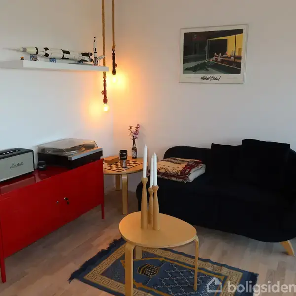 Sort sofa står ved væg med et kunstværk. Rødt skab bærer pladespiller og højttaler. Rundt bord med lys og tæppe foran. Væglampe hænger over hylde.